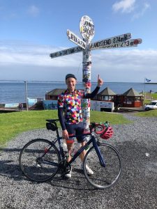 man-with-bicycle-points-to-lands-end-sign-on-his-charity-cycle-for-the-david-nott-foundation